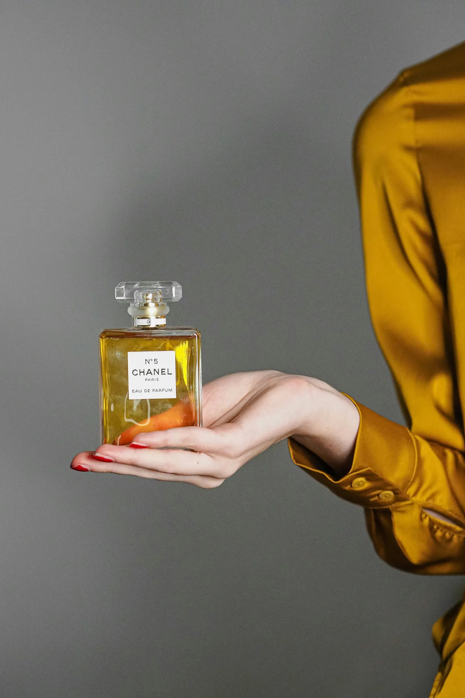 A hand offering a golden flacon against a grey wall, gold sleeve at the edge
